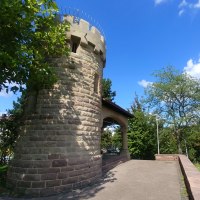 Runder Steinturm mit Zinnen, umgeben von B&auml;umen und blauem Himmel. Der Turm steht auf einer gepflasterten Fl&auml;che., &copy; SMG