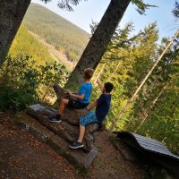 Zwei Kinder sitzen auf einer Holzskulptur im Wald und schauen in die Ferne. Umgeben von B&auml;umen, mit Blick auf eine bewaldete Landschaft., &copy; N&ouml;rdlicher Schwarzwald