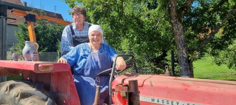 Ein Mann und eine Frau sitzen auf einem roten Traktor im Freien. Die Frau tr&auml;gt ein Kopftuch und Sch&uuml;rze, der Mann eine Brille und Latzhose. Im Hintergrund sind B&auml;ume., &copy; Touristik Bad Wildbad GmbH