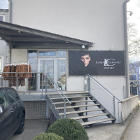 Eingang eines Luisa Cerano Outlets mit Treppe, Werbebanner und Kleidung auf einem Ständer. Ein Auto parkt daneben, Büsche im Vordergrund., © L. Besmer