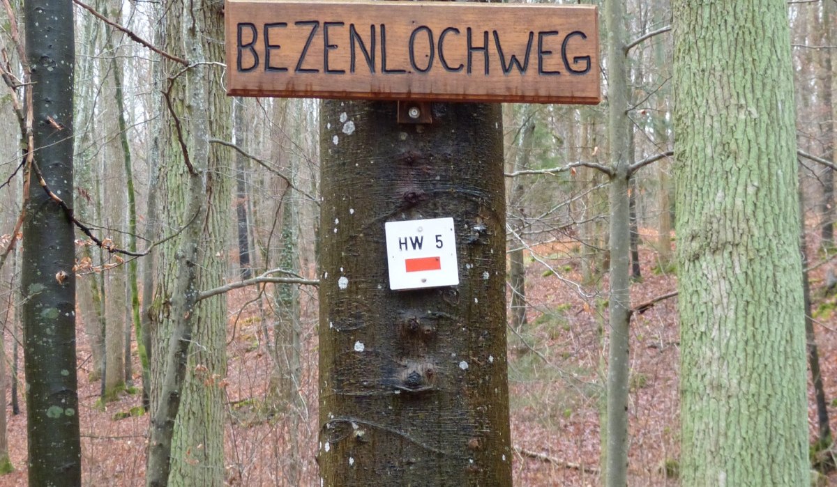 Ein Holzschild mit der Aufschrift 'BEZENLOCHWEG' hängt an einem Baum im Wald. Darunter sind zwei Wandermarkierungen angebracht., © Natur.Nah. Schönbuch & Heckengäu