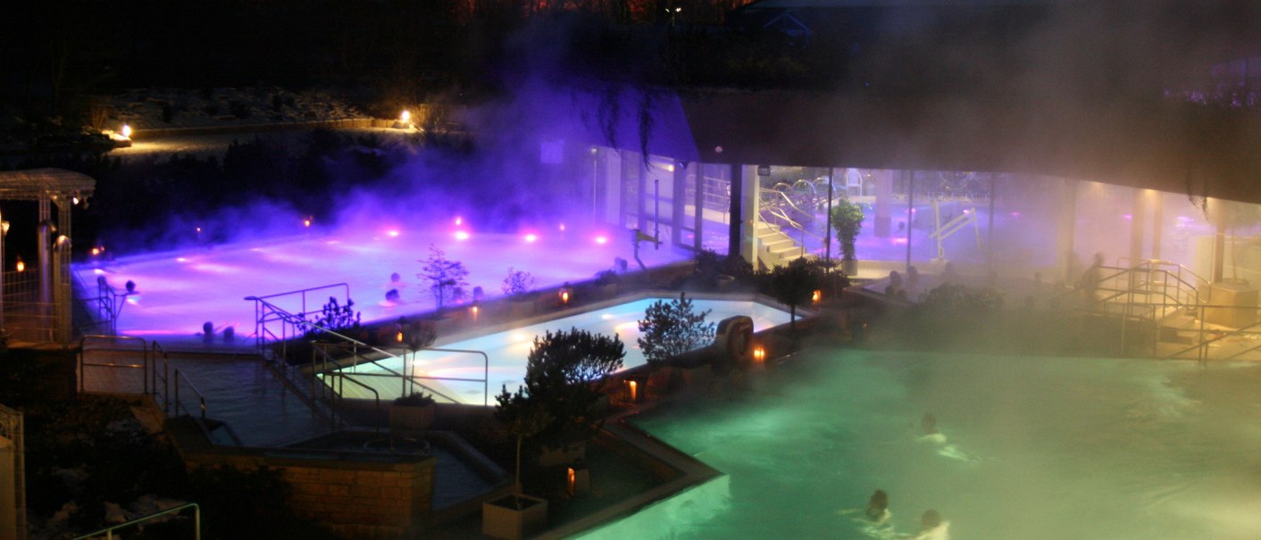 Beleuchtete Außenpools der Panorama Therme Beuren bei Nacht, umgeben von Dampf und Pflanzen. Menschen entspannen im warmen Wasser., © Stuttgart-Marketing GmbH Beleuchtete Außenpools der Panorama Therme Beuren bei Nacht, umgeben von Dampf und Pflanzen. Menschen entspannen im warmen Wasser., © Stuttgart-Marketing GmbH