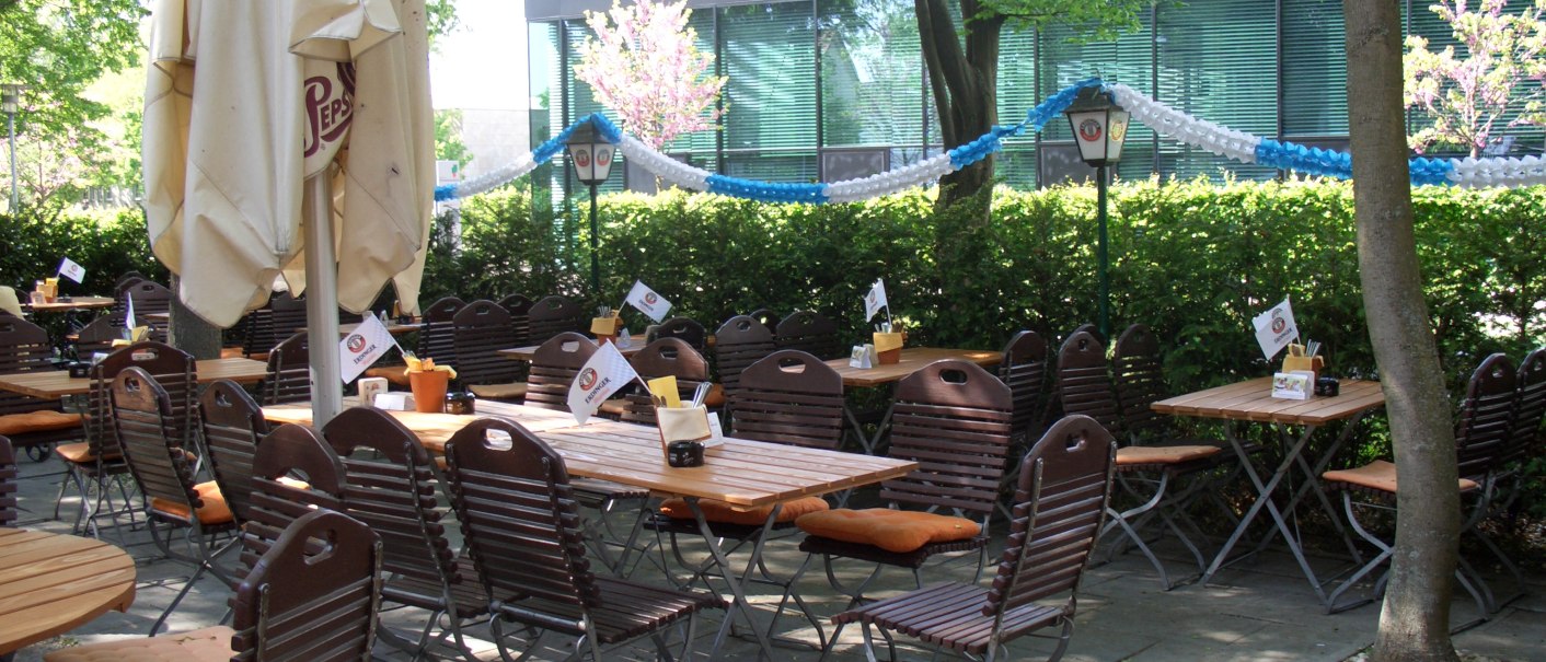 Ein leerer Biergarten mit Holztischen und Stühlen, umgeben von Bäumen und blau-weißen Girlanden., © Wirtshaus zom Schiller Fellbach Ein leerer Biergarten mit Holztischen und Stühlen, umgeben von Bäumen und blau-weißen Girlanden., © Wirtshaus zom Schiller Fellbach