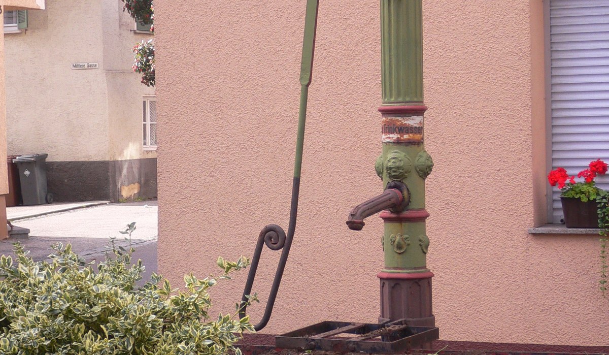Ein grüner, dekorativer Pump-Brunnen steht vor einer rosa Hauswand. Im Vordergrund sind Pflanzen zu sehen., © Land der 1000 Hügel - Kraichgau-Stromberg Ein grüner, dekorativer Pump-Brunnen steht vor einer rosa Hauswand. Im Vordergrund sind Pflanzen zu sehen., © Land der 1000 Hügel - Kraichgau-Stromberg