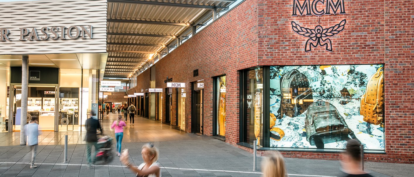 Einkaufsstraße in der Outletcity Metzingen mit Geschäften wie MCM. Menschen schlendern vorbei, einige sind in Bewegung unscharf., © OUTLETCITY AG