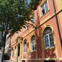 Historisches Gebäude mit roter Fassade und gelben Fensterrahmen, flankiert von Bäumen. Die Sonne wirft Schatten auf den Gehweg., © TELB Historisches Gebäude mit roter Fassade und gelben Fensterrahmen, flankiert von Bäumen. Die Sonne wirft Schatten auf den Gehweg., © TELB