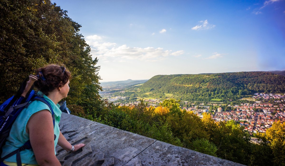 Eine Frau mit Rucksack blickt von einem Aussichtspunkt auf eine bewaldete Hügellandschaft und eine Stadt im Tal. Der Himmel ist klar und sonnig., © Stadt Geislingen an der Steige Eine Frau mit Rucksack blickt von einem Aussichtspunkt auf eine bewaldete Hügellandschaft und eine Stadt im Tal. Der Himmel ist klar und sonnig., © Stadt Geislingen an der Steige