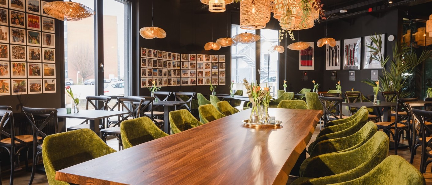 Ein Café mit grünen Samtstühlen, Holztischen und dekorativen Hängelampen. An den Wänden hängen viele Fotos., © TOMAS Ein Café mit grünen Samtstühlen, Holztischen und dekorativen Hängelampen. An den Wänden hängen viele Fotos., © TOMAS