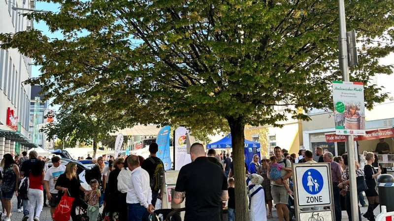 Menschen versammeln sich bei einem Stra&szlig;enfest unter einem gro&szlig;en Baum. Im Hintergrund sind St&auml;nde und ein Schild f&uuml;r eine Fu&szlig;g&auml;ngerzone zu sehen.