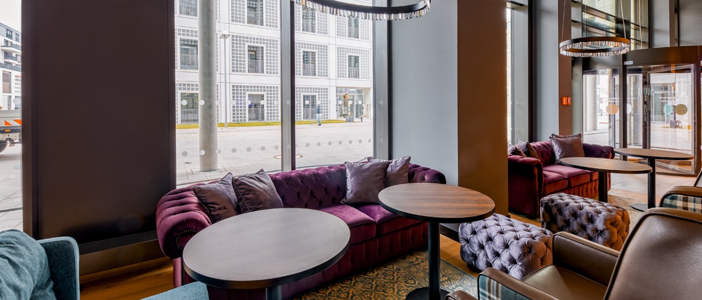 Moderne Lobby mit lila Sofas, runden Tischen und großen Fenstern, die auf eine Straße blicken. Elegante Beleuchtung und gemütliche Atmosphäre., © Premier Inn