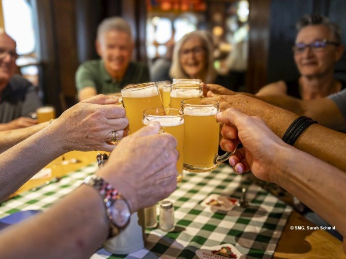 Eine Gruppe von Menschen stößt mit Biergläsern an einem Tisch an. Im Hintergrund sind lächelnde Gesichter zu sehen., © Stuttgart Marketing GmbH Eine Gruppe von Menschen stößt mit Biergläsern an einem Tisch an. Im Hintergrund sind lächelnde Gesichter zu sehen., © Stuttgart Marketing GmbH