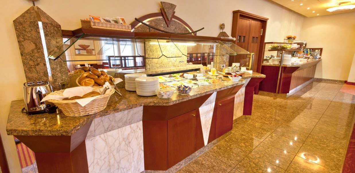 Ein Frühstücksbuffet in einem Hotel mit einer Auswahl an Brötchen, Aufschnitt, Obst und Getränken auf einer langen Theke., © Novum Hospitality