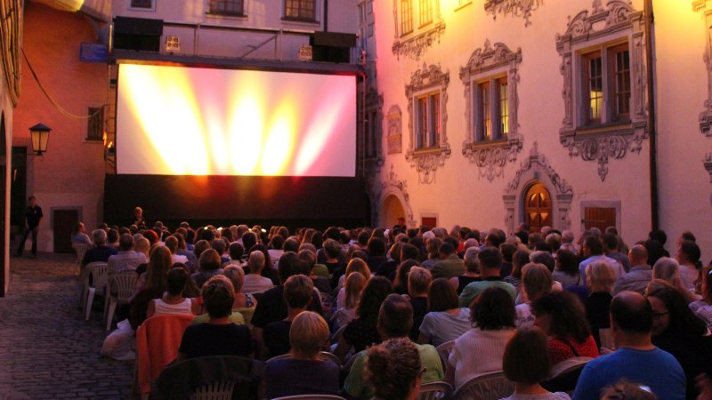 Mit mehreren Lichtern angestrahlte große Filmleinwand im Schlossinnenhof, davor Zuschauer, Bild in Dämmerlicht aufgenommen, © Sonnenlichtspiele e.V.