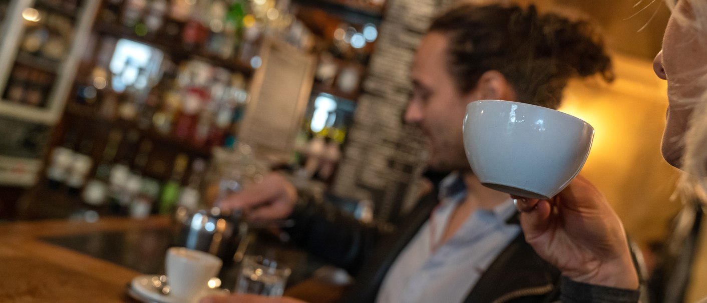 Zwei Personen in einem Café. Eine Person trinkt aus einer Tasse, während die andere Kaffee zubereitet. Im Hintergrund sind Regale mit Flaschen zu sehen., © Stuttgart Marketing GmbH, Fotografin Martina Denker Zwei Personen in einem Café. Eine Person trinkt aus einer Tasse, während die andere Kaffee zubereitet. Im Hintergrund sind Regale mit Flaschen zu sehen., © Stuttgart Marketing GmbH, Fotografin Martina Denker