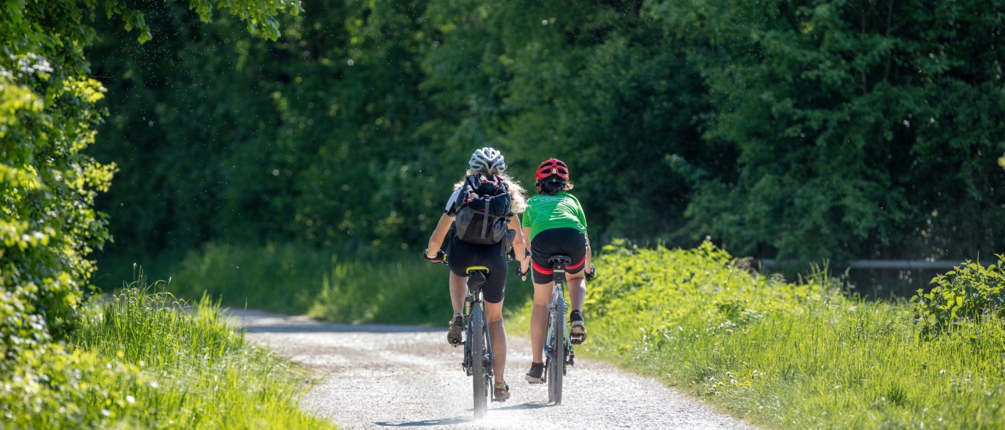Zwei Radfahrer fahren auf einem sonnigen Waldweg. Sie tragen Helme und Rucks&auml;cke, umgeben von gr&uuml;ner Natur., &copy; SMG, Achim Mende