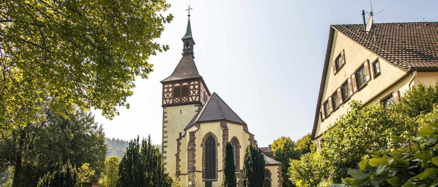 Kirche in Bad Liebenzell mit Fachwerkturm, umgeben von Bäumen und einem Wohngebäude bei sonnigem Wetter., © Stuttgart-Marketing GmbH, Sarah Schmid