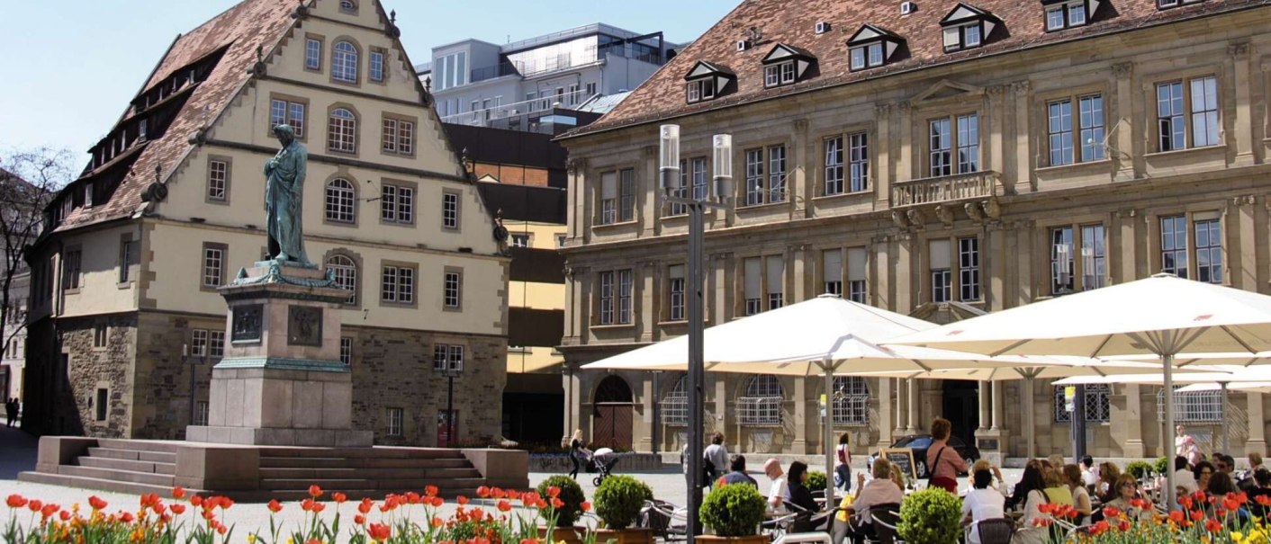 Der Schillerplatz in Stuttgart mit einer Statue im Vordergrund, umgeben von historischen Geb&auml;uden und Menschen, die unter Sonnenschirmen sitzen., &copy; Stuttgart-Marketing GmbH