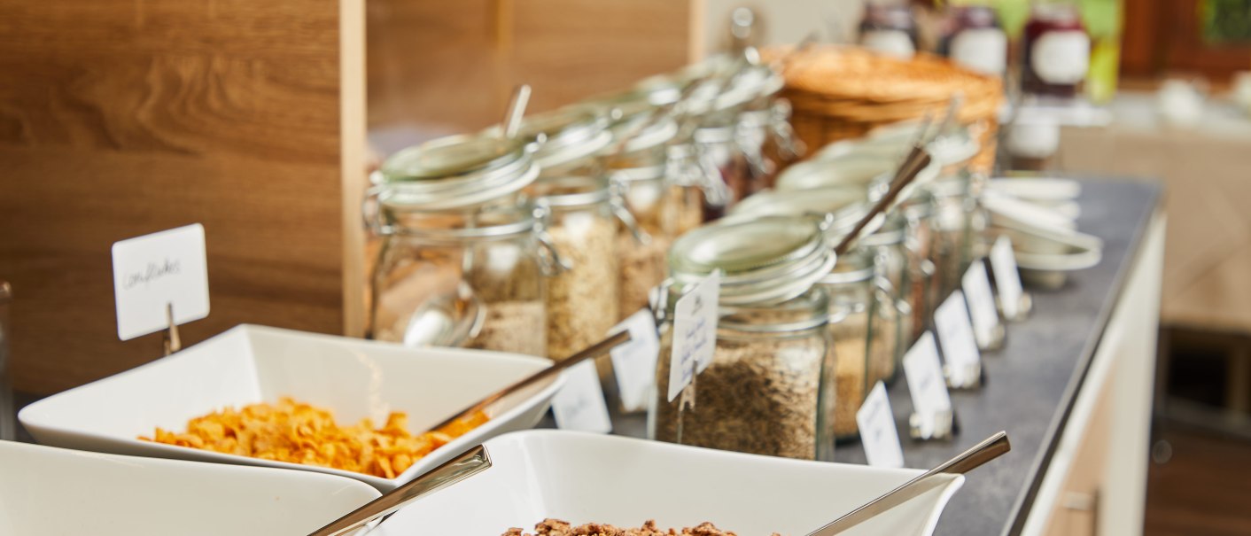 Frühstücksbuffet mit Müsli-Schalen und Gläsern voller Zutaten. Verschiedene Cerealien und Körner sind in Gläsern aufgereiht., © TOMAS