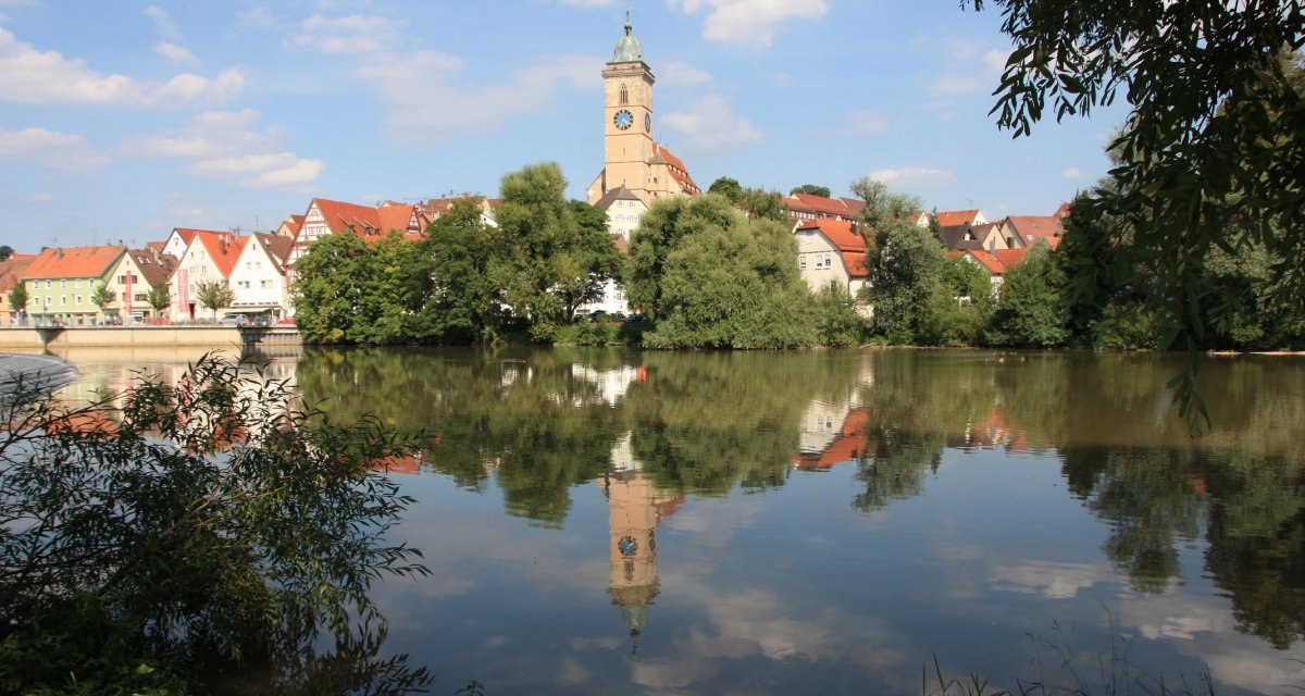 Panorama von Nürtingen mit Kirche und Fachwerkhäusern, die sich im ruhigen Wasser spiegeln, umgeben von Bäumen und blauem Himmel., © Stadt Nürtingen Panorama von Nürtingen mit Kirche und Fachwerkhäusern, die sich im ruhigen Wasser spiegeln, umgeben von Bäumen und blauem Himmel., © Stadt Nürtingen