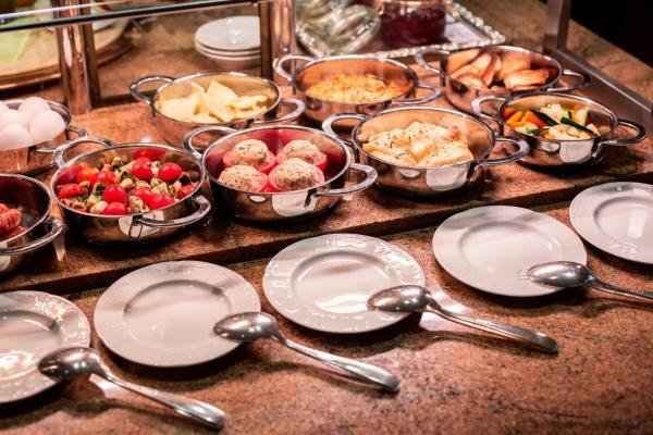 Frühstücksbuffet mit Metallschalen voller Speisen, darunter Tomaten und Brötchen. Leere Teller und Löffel sind davor platziert., © Kronen Hotel GmbH