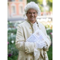 Ein Mann in barocker Kleidung und wei&szlig;er Per&uuml;cke steht l&auml;chelnd in einem Garten und h&auml;lt ein Buch. Im Hintergrund sind Blumen und ein Geb&auml;ude zu sehen., &copy; Touristik und Marketing GmbH