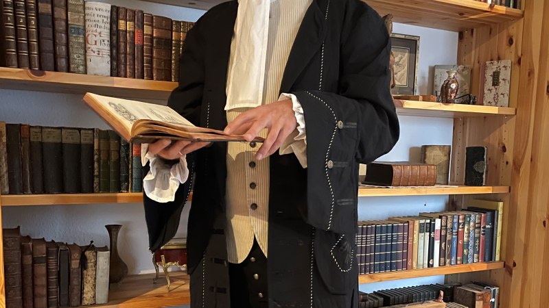 Eine Person in historischer Kleidung mit Dreispitz und R&uuml;schenhemd steht vor einem B&uuml;cherregal und h&auml;lt ein aufgeschlagenes Buch., &copy; Gerd Jenner