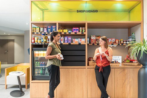 Zwei Frauen unterhalten sich vor einem kleinen Hotelshop mit Snacks und Getränken. Der Shop trägt das Schild 'SHOP@ADAGIO'., © adagio access Stuttgart Airport Messe