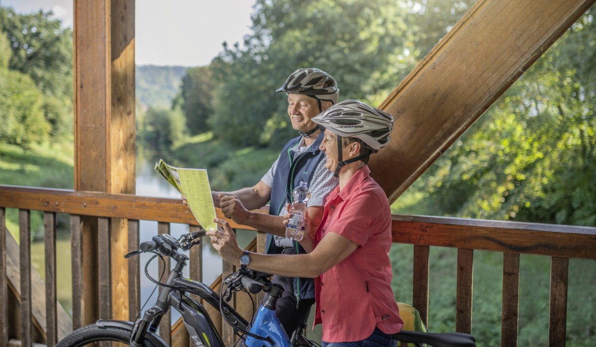 Zwei Radfahrer mit Helmen stehen auf einer Holzbrücke und betrachten eine Karte. Im Hintergrund ist grüne Landschaft zu sehen., © Rems-Murr-Kreis, Jan Bürgermeister