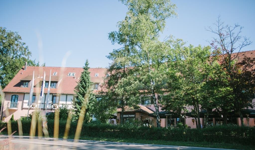 Ein Hotel mit rotem Dach, umgeben von Bäumen, unter blauem Himmel. Im Vordergrund sind Fahnen und eine Straße zu sehen., © relexa Waldhotel Schatten Ein Hotel mit rotem Dach, umgeben von Bäumen, unter blauem Himmel. Im Vordergrund sind Fahnen und eine Straße zu sehen., © relexa Waldhotel Schatten