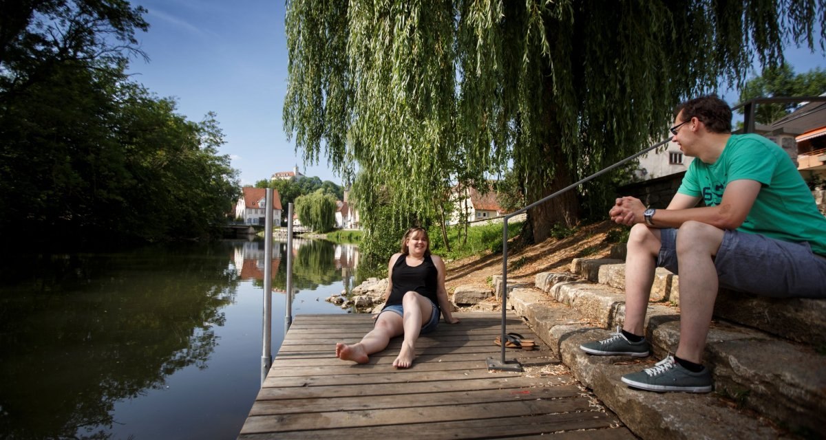Zwei Personen sitzen entspannt auf einem Holzsteg am Flussufer. Eine Frau sitzt barfuß, ein Mann sitzt auf Steinstufen. Im Hintergrund sind Bäume und Häuser., © Stoppel Zwei Personen sitzen entspannt auf einem Holzsteg am Flussufer. Eine Frau sitzt barfuß, ein Mann sitzt auf Steinstufen. Im Hintergrund sind Bäume und Häuser., © Stoppel
