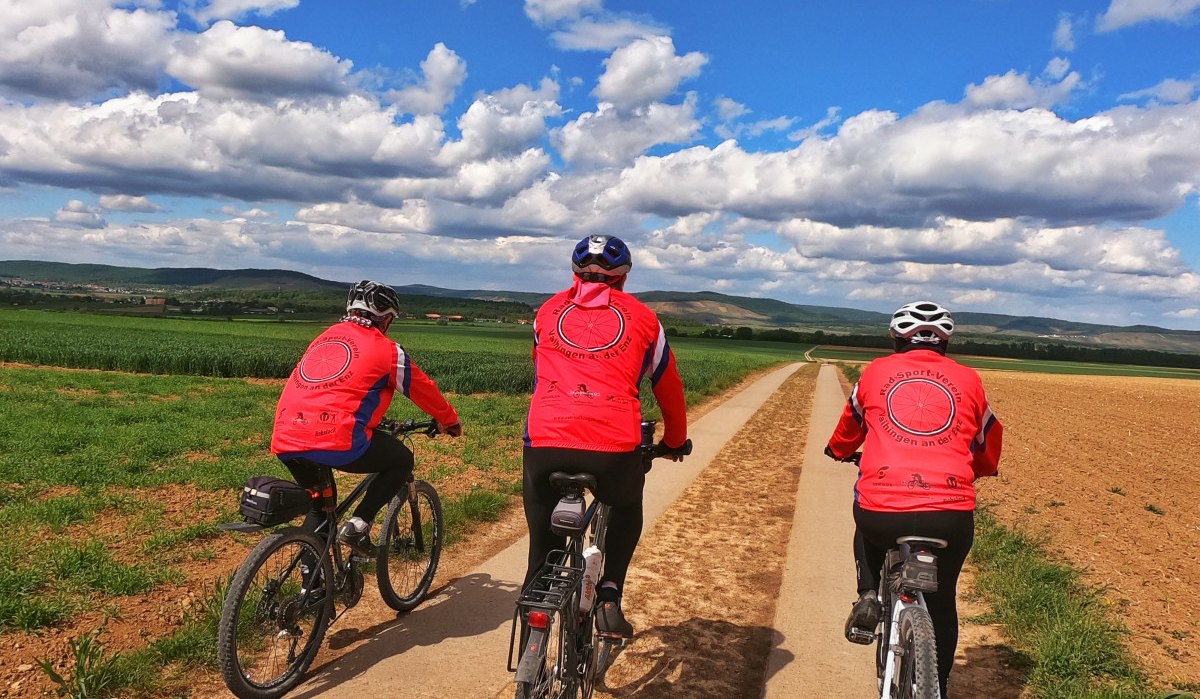 Drei Radfahrer in roten Trikots fahren auf einem schmalen Feldweg durch eine ländliche Landschaft unter blauem Himmel mit Wolken., © Land der 1000 Hügel - Kraichgau-Stromberg Drei Radfahrer in roten Trikots fahren auf einem schmalen Feldweg durch eine ländliche Landschaft unter blauem Himmel mit Wolken., © Land der 1000 Hügel - Kraichgau-Stromberg