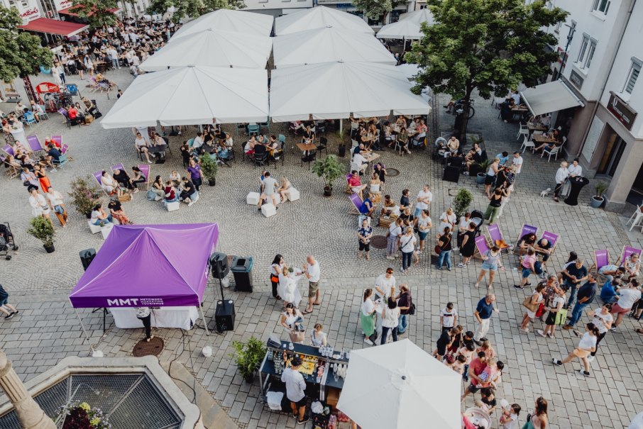 Belebter Platz in Metzingen mit Menschen unter weißen Sonnenschirmen und einem lila Zelt. Viele Besucher genießen die entspannte Atmosphäre.