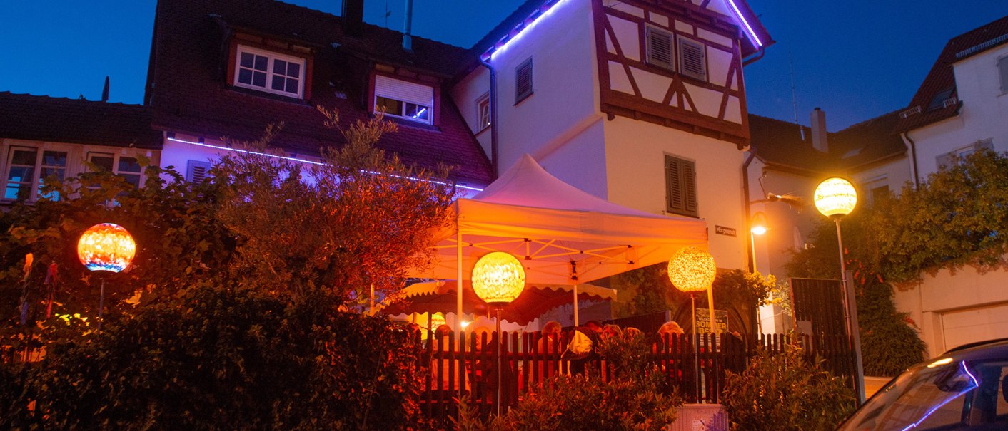 Ein beleuchtetes Fachwerkhaus mit einem weißen Zelt im Garten. Bunte Laternen und Lichterketten schaffen eine gemütliche Atmosphäre bei Nacht., © Weingut Ruoff