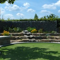 Minigolfplatz mit künstlichem Grün, umgeben von Pflanzen und Steinmauern. Ein Baum spendet Schatten, der Himmel ist blau mit weißen Wolken., © Anja Beyerle & Ralf Bähr GbR