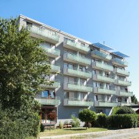 Ein modernes, mehrstöckiges Wohngebäude mit Balkonen und Glasgeländern, umgeben von Bäumen und einem klaren blauen Himmel., © TOMAS