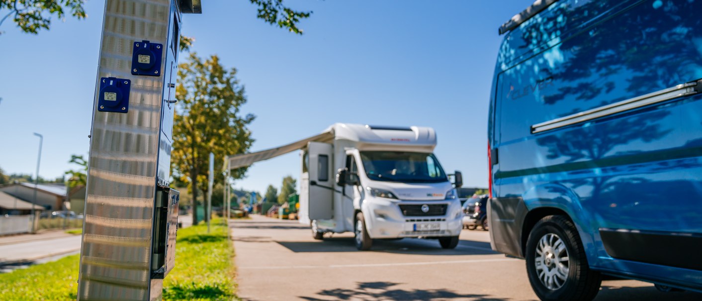 Wohnmobilstellplatz mit einem weißen Wohnmobil und einem blauen Van. Im Vordergrund eine Stromsäule, im Hintergrund Bäume und blauer Himmel., © Stuttgart-Marketing GmbH, Thomas Niedermüller