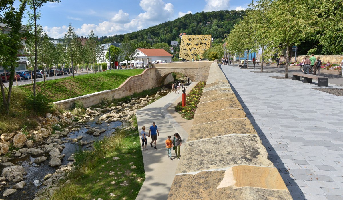 Fußweg entlang eines kleinen Flusses, gesäumt von Bäumen und Blumen. Moderne Architektur im Hintergrund, sonniges Wetter., © Burkhard Walther Fußweg entlang eines kleinen Flusses, gesäumt von Bäumen und Blumen. Moderne Architektur im Hintergrund, sonniges Wetter., © Burkhard Walther