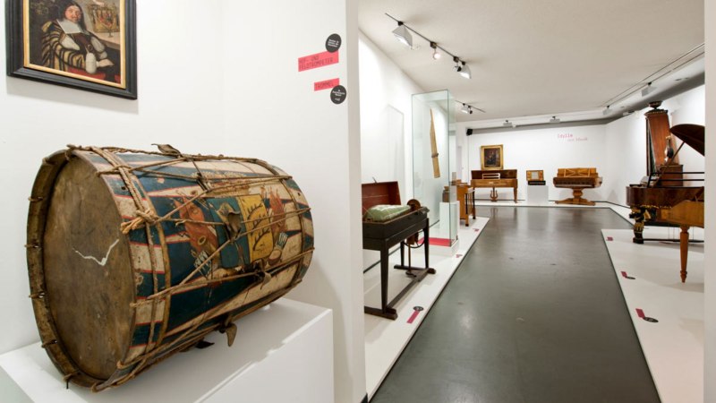 Museumsausstellung mit historischen Musikinstrumenten, darunter eine bemalte Trommel im Vordergrund und verschiedene Tasteninstrumente im Hintergrund., &copy; Landesmuseum W&uuml;rttemberg