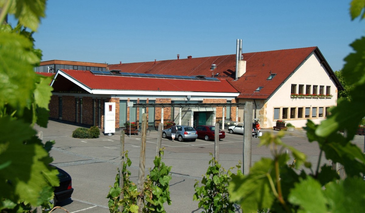 Gebäude der Fellbacher Weingärtner mit rotem Dach, umgeben von Weinreben. Autos parken auf dem Vorplatz. Klare, sonnige Wetterbedingungen., © Remstal Tourismus e.V. Gebäude der Fellbacher Weingärtner mit rotem Dach, umgeben von Weinreben. Autos parken auf dem Vorplatz. Klare, sonnige Wetterbedingungen., © Remstal Tourismus e.V.