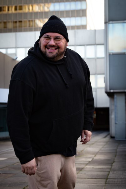 Ein Mann in schwarzem Hoodie und Mütze lächelt in einer städtischen Umgebung mit modernen Gebäuden im Hintergrund., © SBEntertainment GmbH & Co. KG Ein Mann in schwarzem Hoodie und Mütze lächelt in einer städtischen Umgebung mit modernen Gebäuden im Hintergrund., © SBEntertainment GmbH & Co. KG