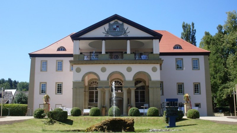 Schloss D&auml;tzingen mit symmetrischer Fassade, rotem Dach und einem Brunnen im Vordergrund. Der Himmel ist klar und blau., &copy; Natur.Nah. Sch&ouml;nbuch & Heckeng&auml;u