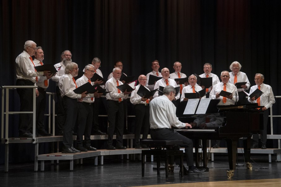 Ein M&auml;nnerchor in wei&szlig;en Hemden und orangefarbenen Krawatten singt auf einer B&uuml;hne. Ein Pianist begleitet sie am Fl&uuml;gel.