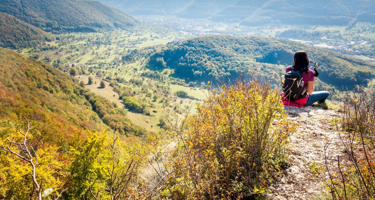 Eine Person sitzt auf einem Hügel und blickt ins Lenninger Tal. Die Landschaft ist von bewaldeten Hügeln und weiten Wiesen geprägt., © hochgehberge