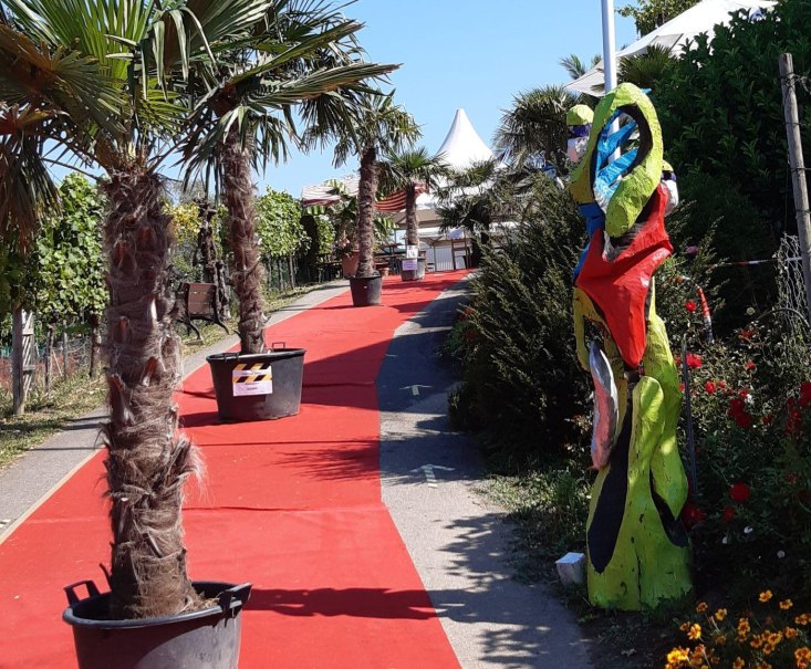 Ein roter Teppich f&uuml;hrt durch einen Garten mit Palmen in T&ouml;pfen und bunten Skulpturen. Im Hintergrund sind wei&szlig;e Zelte zu sehen., &copy; Weingut Bayer