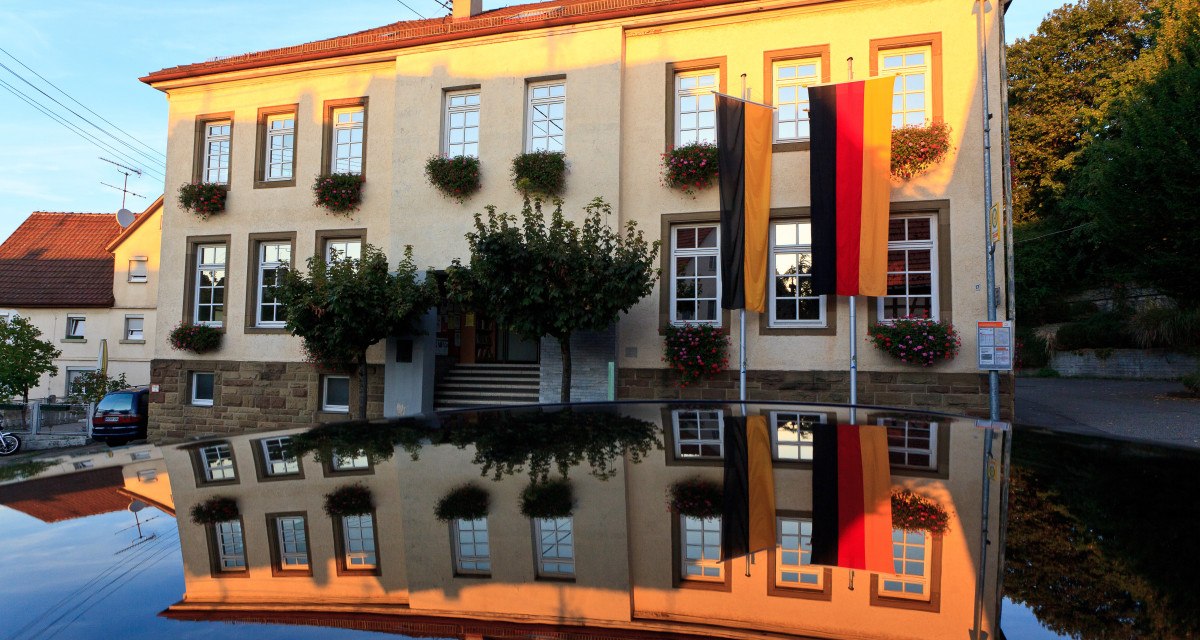 Das Rathaus in Waiblingen-Hohenacker, geschmückt mit Blumen und deutschen Flaggen, spiegelt sich auf einer glänzenden Oberfläche wider., © Waiblingen - Stuttgart-Marketing GmbH Das Rathaus in Waiblingen-Hohenacker, geschmückt mit Blumen und deutschen Flaggen, spiegelt sich auf einer glänzenden Oberfläche wider., © Waiblingen - Stuttgart-Marketing GmbH