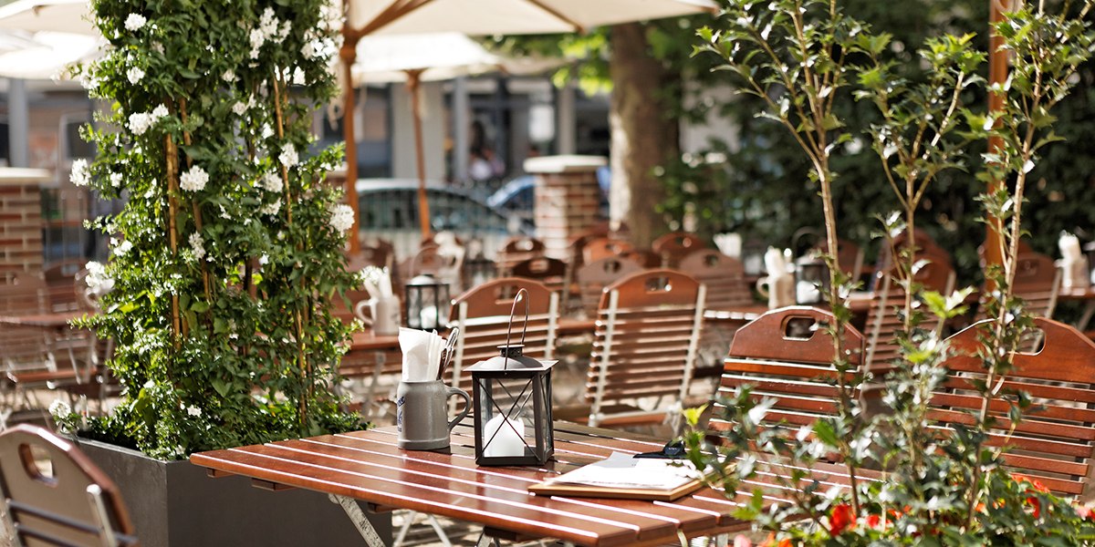 Ein gemütlicher Biergarten mit Holztischen und -stühlen, umgeben von grünen Pflanzen und Sonnenschirmen. Eine Laterne steht auf dem Tisch., © Parkhotel Stuttgart Messe-Airport