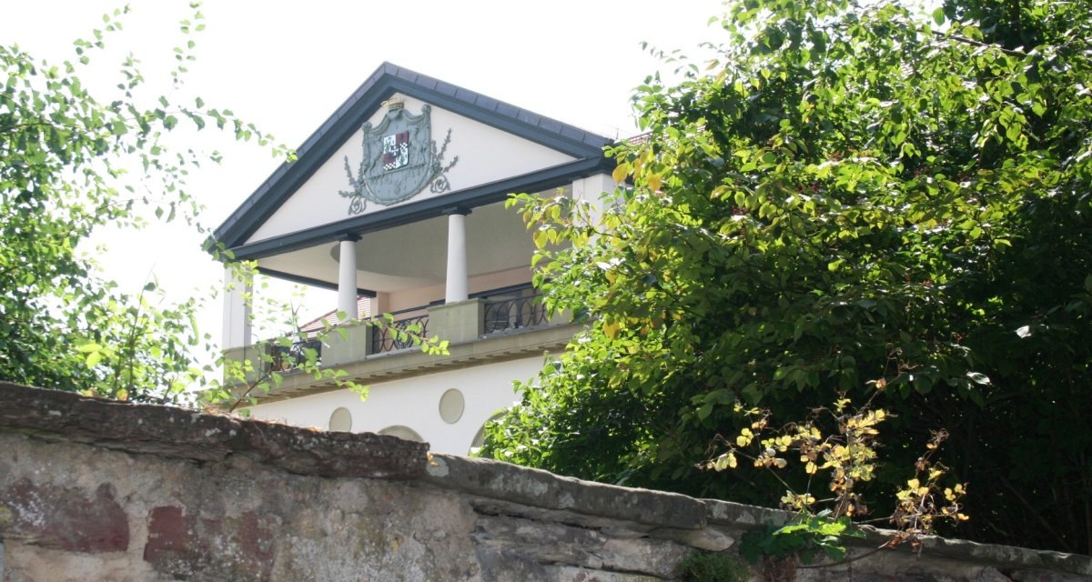 Ein Gebäude mit Säulen und einem Wappen auf der Fassade, umgeben von Bäumen und einer Steinmauer im Vordergrund., © Natur.Nah. Schönbuch & Heckengäu Ein Gebäude mit Säulen und einem Wappen auf der Fassade, umgeben von Bäumen und einer Steinmauer im Vordergrund., © Natur.Nah. Schönbuch & Heckengäu