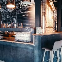 Moderne Bar mit industriellem Design, hohen Stühlen und Wandkunst. Gläser stehen auf der Theke, im Hintergrund eine Tafel mit Getränken., © Kraftpaule Moderne Bar mit industriellem Design, hohen Stühlen und Wandkunst. Gläser stehen auf der Theke, im Hintergrund eine Tafel mit Getränken., © Kraftpaule