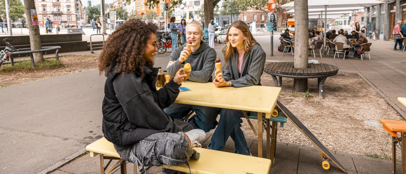 Drei Personen sitzen an einem gelben Tisch im Freien und essen Eis. Im Hintergrund sind Bäume, Fahrräder und ein Café zu sehen., © Stuttgart Marketing GmbH, Sarah Schmid