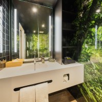 Modernes Badezimmer mit weißem Waschbecken, Spiegel und Dschungelmotiv an der Wand. Schwarze Fliesen und helle Beleuchtung ergänzen das Design., © Rioca Stuttgart Posto 4 GmbH